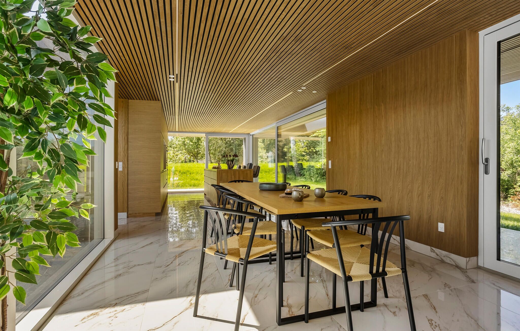 Un espace de restauration moderne avec un plafond en bois, un sol en marbre et une vue sur un jardin grâce à de grandes fenêtres.