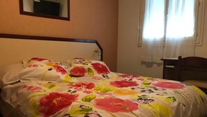 A bedroom with a floral bedspread, a wooden headboard, a bedside table, and a chair. - HOTEL MYOSOTIS (Lourdes)