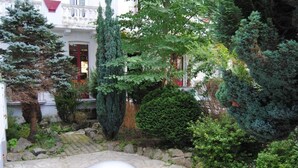 A courtyard with stone flooring, a variety of trees, and a building with a red door in the background. - HOTEL MYOSOTIS (Lourdes)