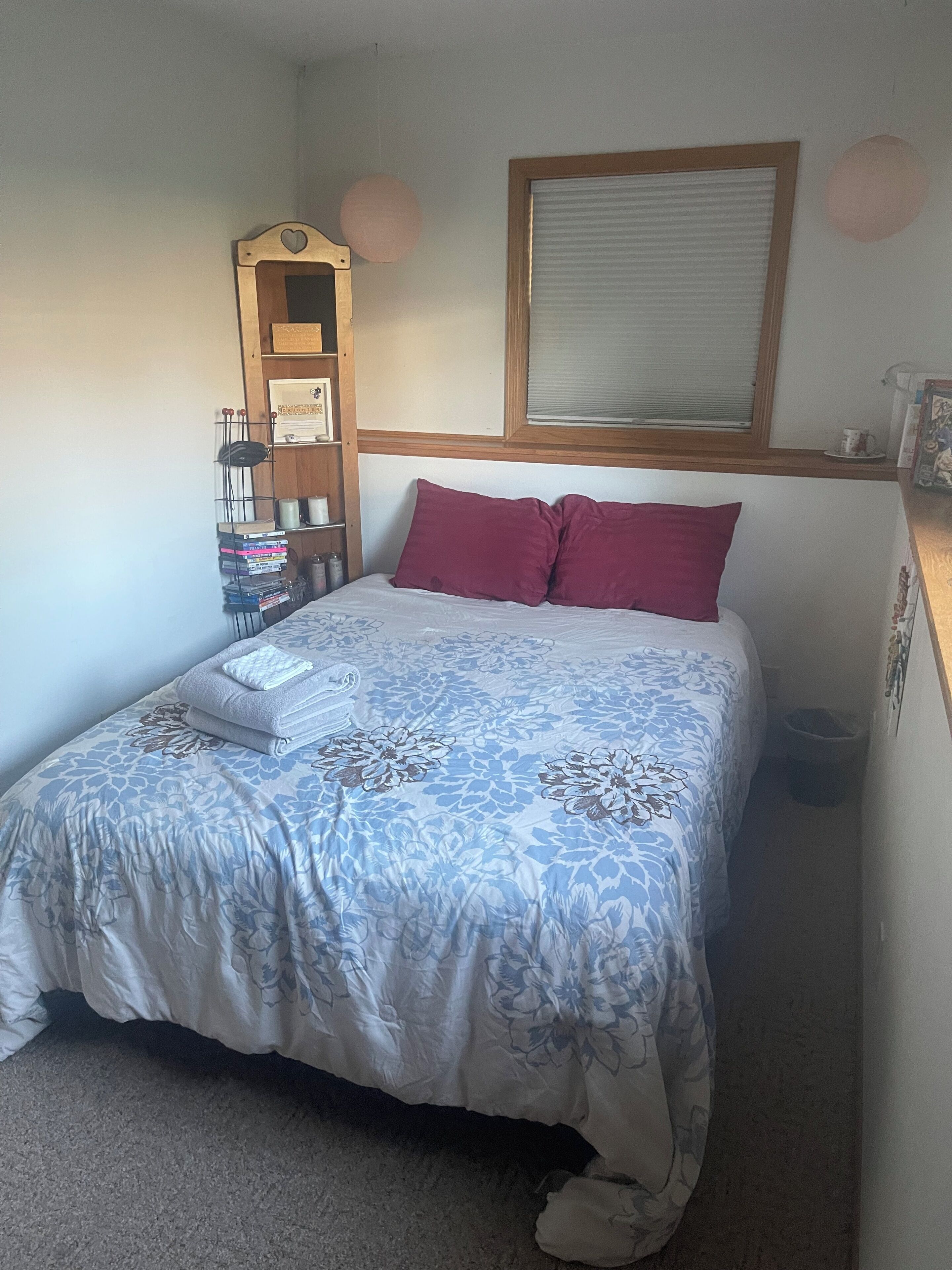 Een slaapkamer met een bed, kussens, een nachtkastje, een raam en een wandplank met boeken.