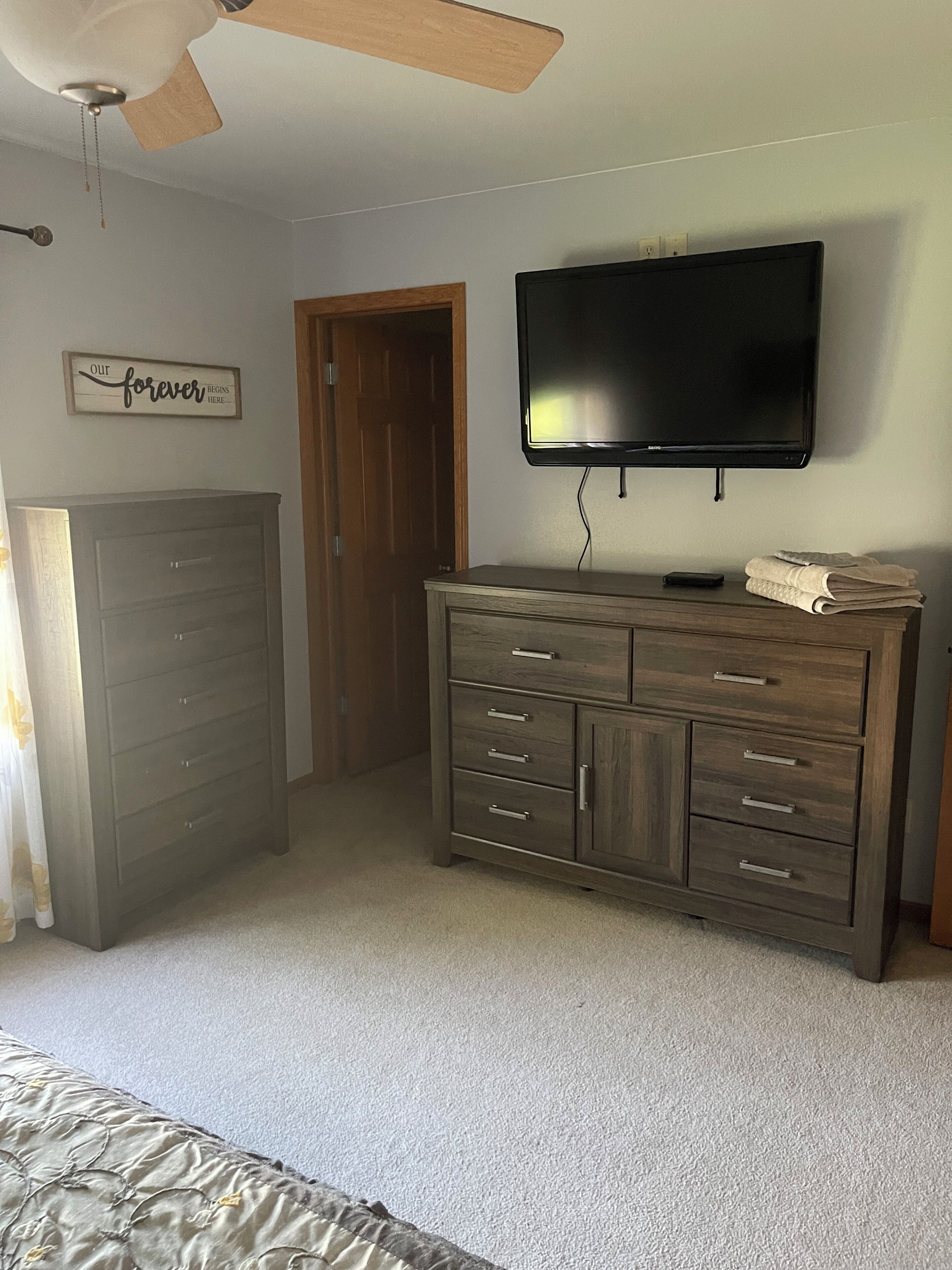 Een slaapkamer met een bed, een dressoir, een televisie, een plafondventilator en een deur.