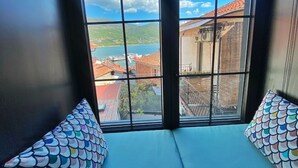 Une pièce dotée d’une grande fenêtre donnant sur un plan d’eau et des bâtiments. On y trouve deux fauteuils bleus avec des coussins aux motifs colorés.