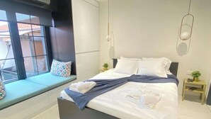 Une chambre moderne avec un grand lit, un coin lecture près de la fenêtre et une table de chevet agrémentée d’une plante.