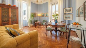 Ein Zimmer mit gestreifter Tapete, einem Holzschrank, einer Couch, einem Esstisch mit Stühlen, einer Kandelaber und gerahmten Bildern an der Wand.