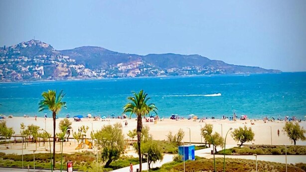 A beach with palm trees, a clear blue sea, and a distant mountain range. - Relaxing Apt Near the Beach (Castello d'Empuries)