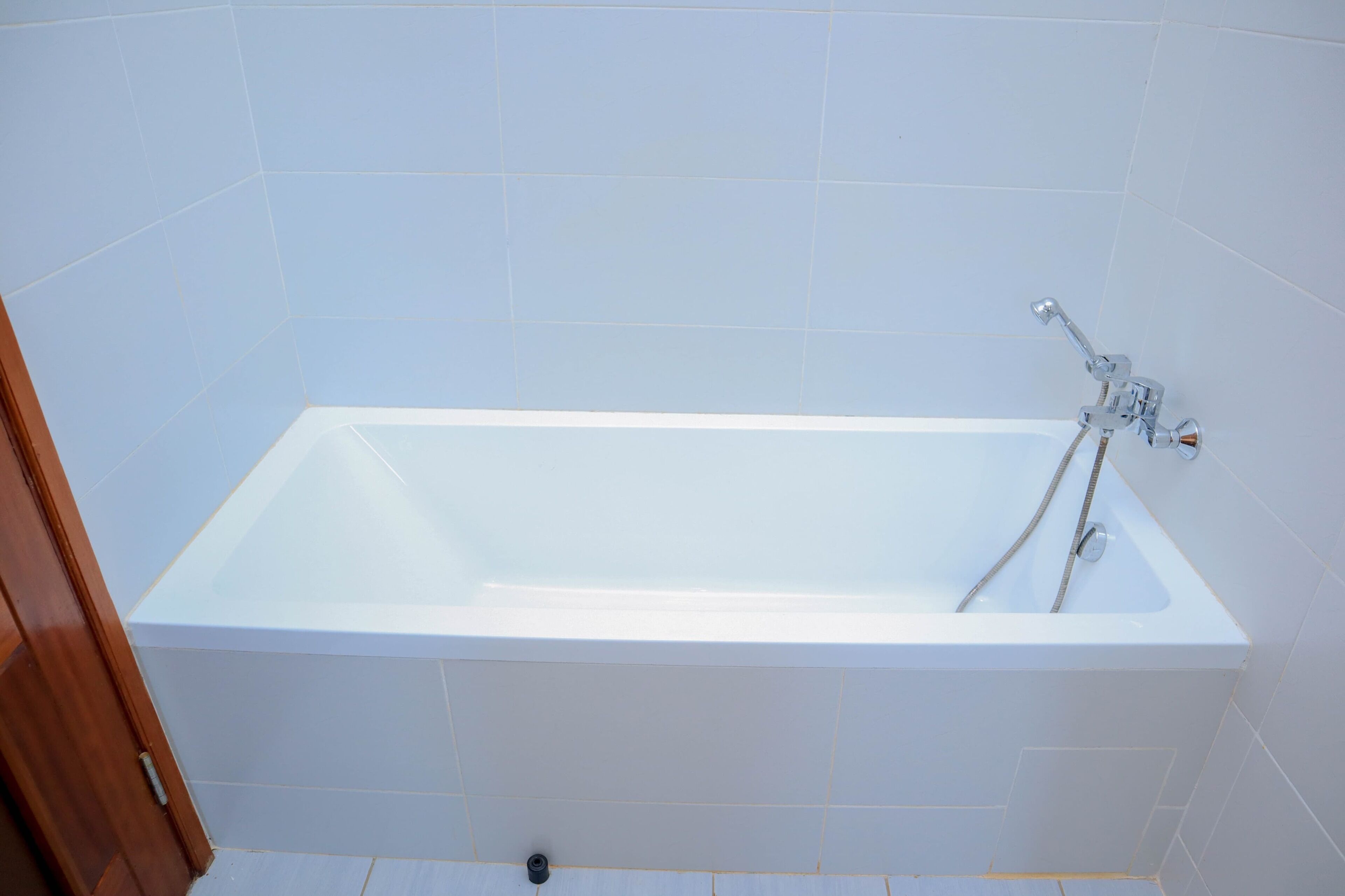 A rectangular bathtub with a chrome faucet and two hoses, set against a tiled wall.