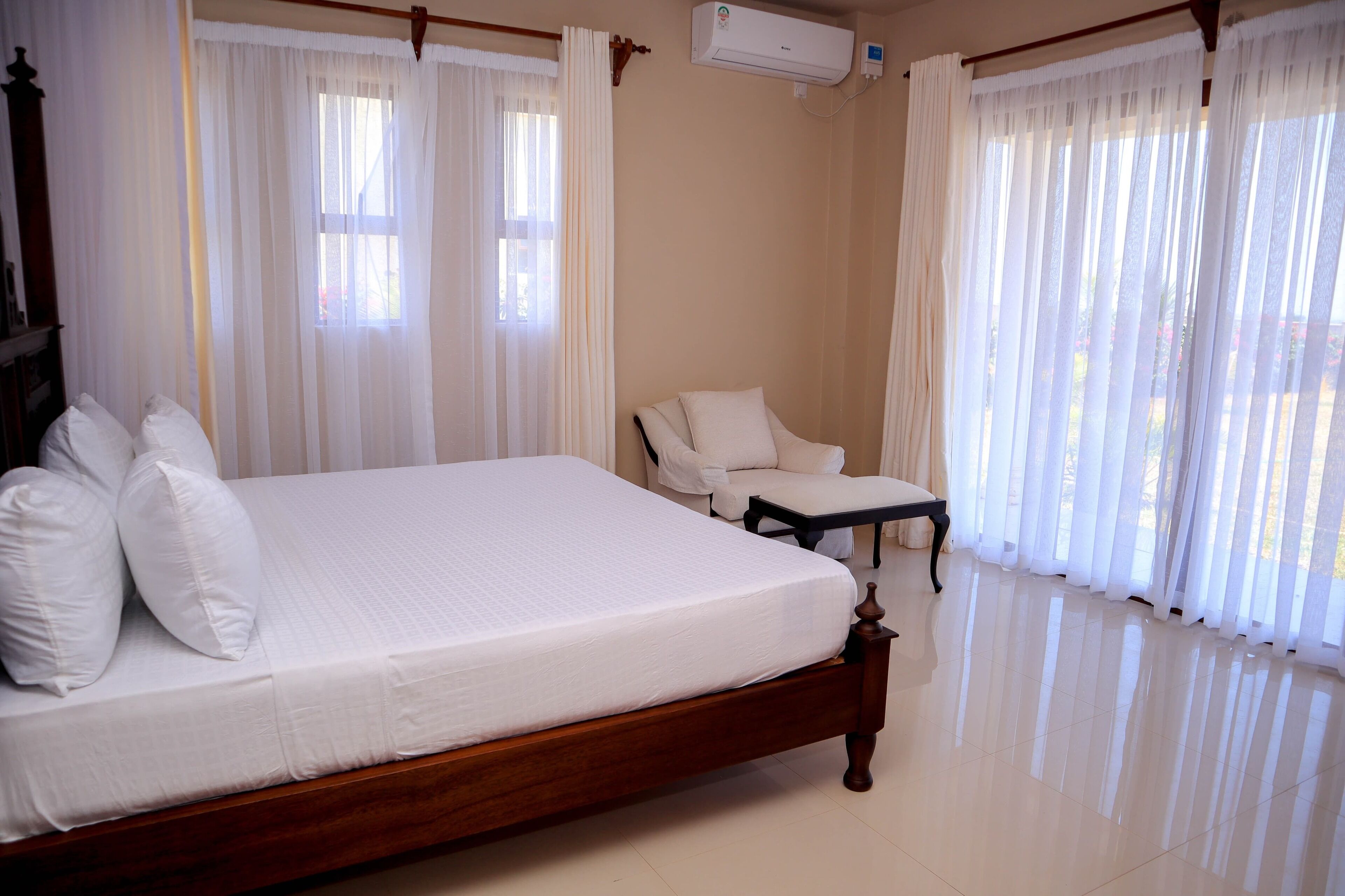 A bedroom with a wooden bed, white bedding, a chair, and sheer curtains.