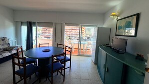 A room with a round table, chairs, a sofa, a TV, and a cabinet. There is a sliding glass door leading to an outdoor area with a view of buildings.