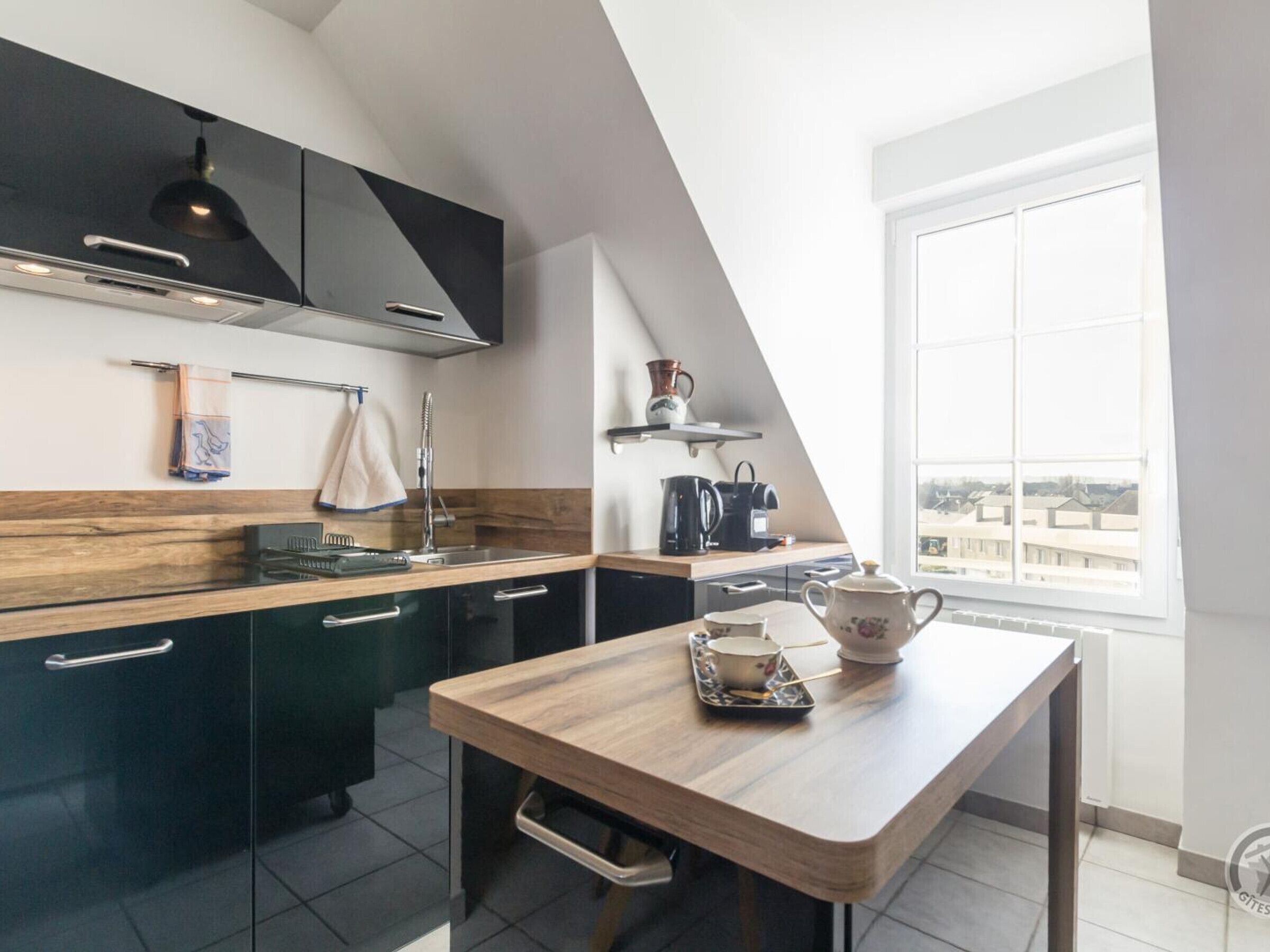 A modern kitchen with dark cabinets, a wooden countertop, and a dining table with a teapot and cup.