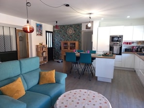 Una sala de estar moderna con un sofá azul, un área de comedor con sillas azules, una cocina con armarios blancos y una pared decorada.