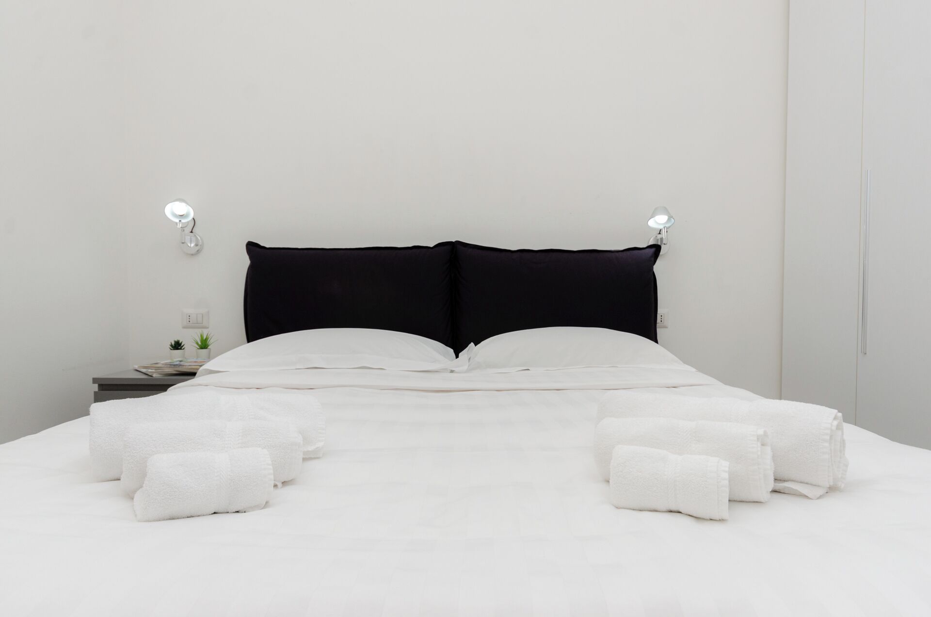 A neatly made bed with white linens and pillows, a bedside table with a lamp, and a small plant on the nightstand.