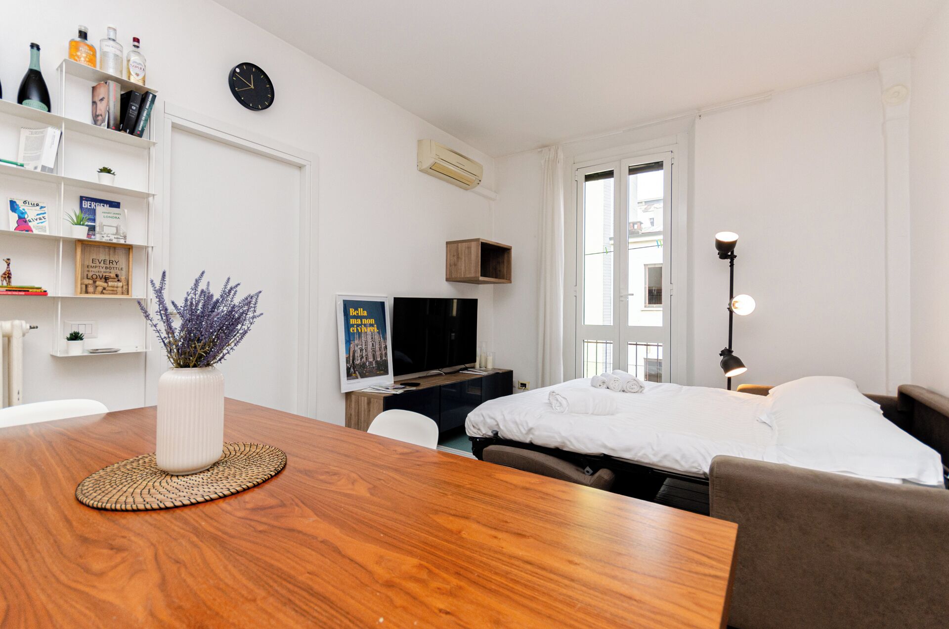 A modern hotel room with a wooden dining table, a bed with white linens, a television, and a large window with blinds.