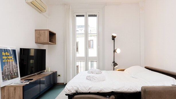 A hotel room with a bed, a TV, a desk with a computer, a lamp, and a window with a view of buildings. - VivimiHome-Stelvio House (Milan)