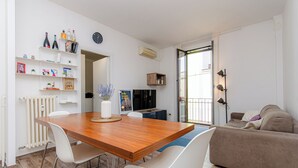 A modern living room with a wooden dining table, white chairs, a sofa, and a TV. There is a bookshelf with books and decorative items, and a radiator on the wall. - VivimiHome-Stelvio House (Milan)