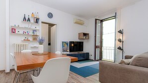 A modern living room with a wooden dining table, white chairs, a flat-screen TV, a sofa, and a large window. - VivimiHome-Stelvio House (Milan)