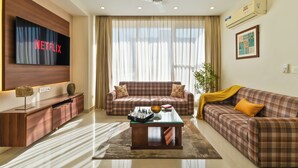 A modern living room with a flat-screen TV, a ceiling fan, and a large window with curtains. - Auro Bliss - An Opulent 3bhk Penthouse in Dehradun (Dehradun)