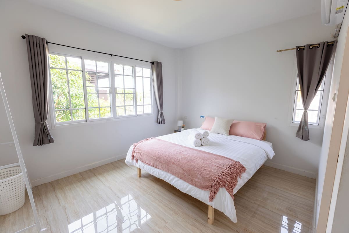A bedroom with a large window, a bed with rolled towels, a bedside table, and a laundry basket.