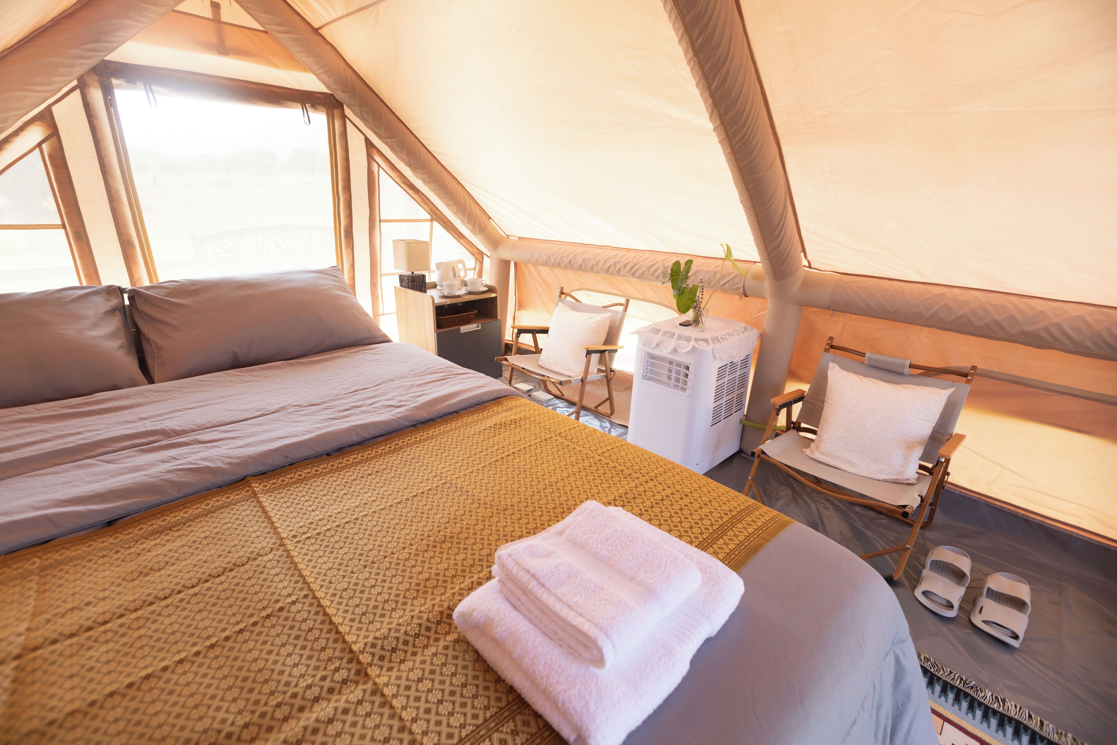 A tented accommodation with a bed, a chair, a table with a lamp, and a small air cooler.