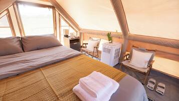 A tented accommodation with a bed, a chair, a table with a lamp, and a small air cooler.