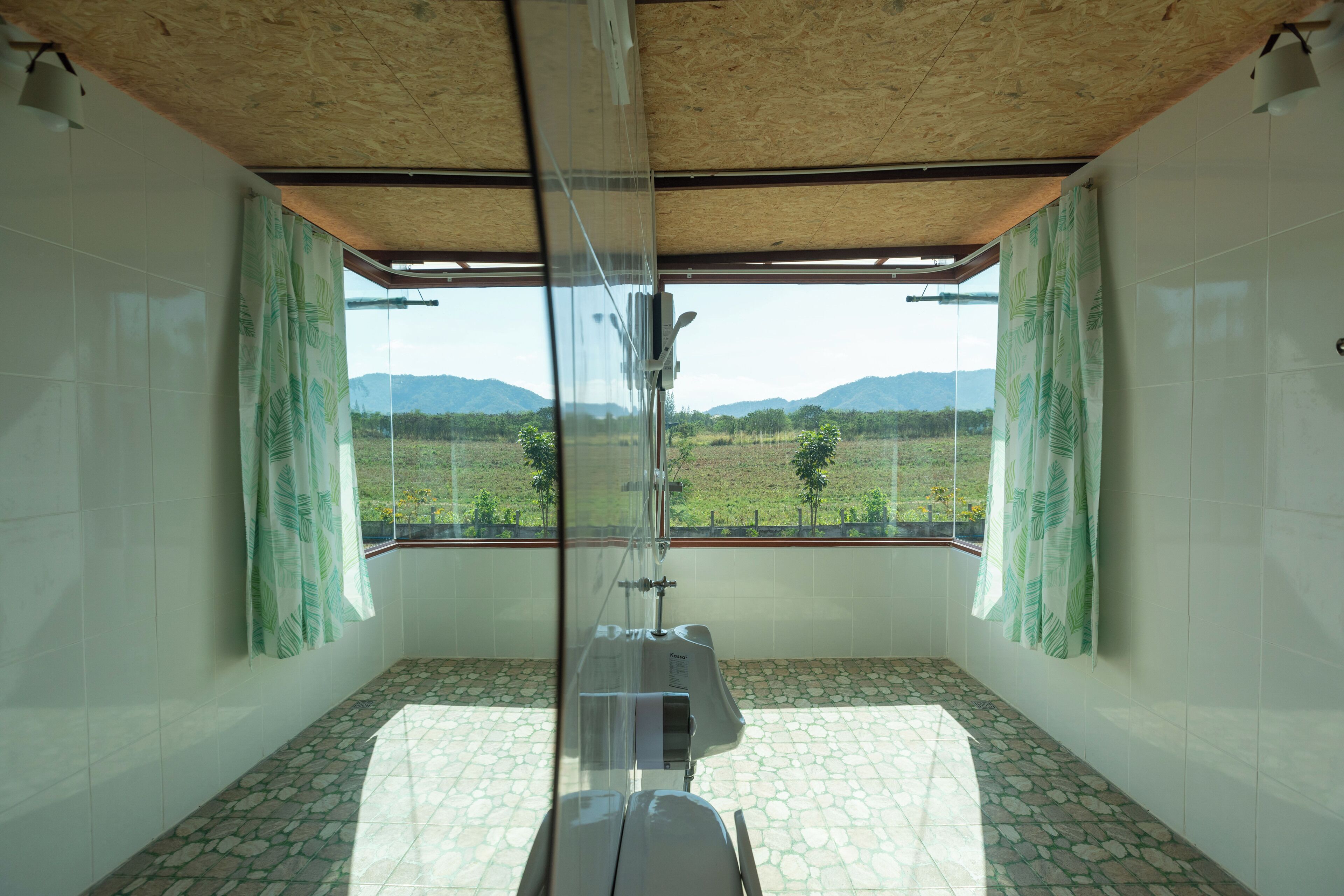 A bathroom with a glass shower enclosure, white tiled walls, and a view of a green landscape through large windows.