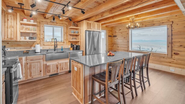 A wooden cabin kitchen with a central island, stainless steel appliances, and a large window offering a view. - Mountain Cabin Near Trails, Skiing & Yellowstone 3 Bedroom Cabin (Belfry)