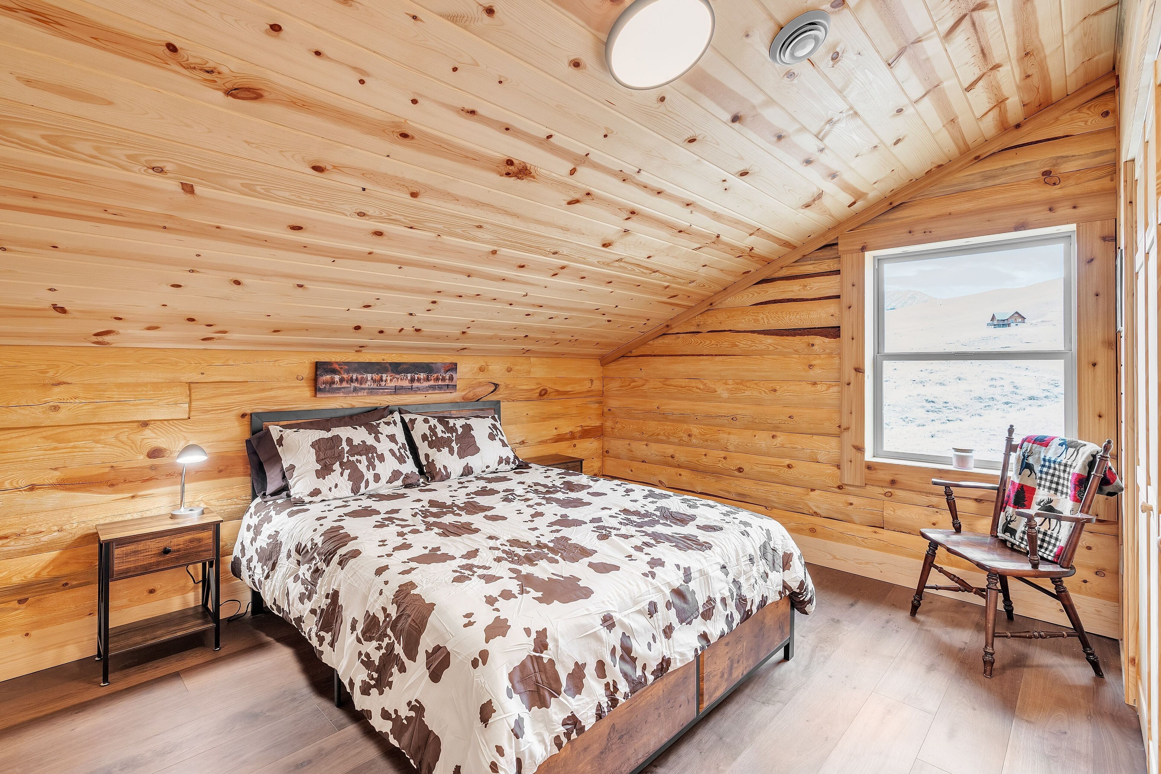 A cozy wooden cabin bedroom with a bed, nightstand, and a window with a view of a snowy landscape.