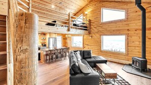 A spacious loft-style room with a large black leather sofa, a wooden dining table with chairs, a kitchen area, and a loft with a staircase. - Mountain Cabin Near Trails, Skiing & Yellowstone 3 Bedroom Cabin (Belfry)