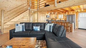 A log cabin interior with a black leather sectional sofa, wooden staircase, and a kitchen area. - Mountain Cabin Near Trails, Skiing & Yellowstone 3 Bedroom Cabin (Belfry)