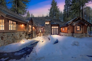 Una cabaña de troncos con paredes y cimientos de piedra, rodeada de nieve y árboles.