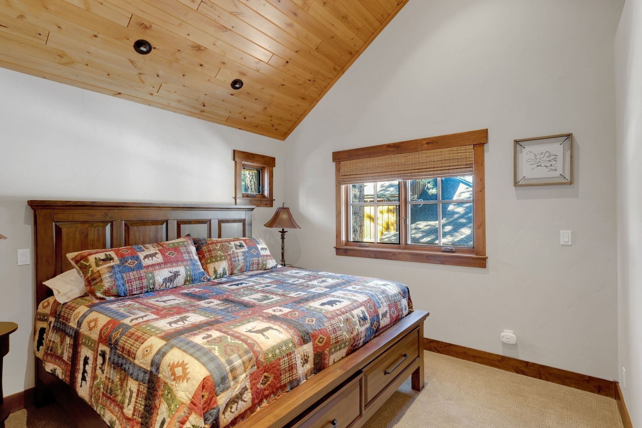 Un dormitorio con techo de madera, una cama grande con colcha colorida, una ventana con persianas y un cuadro enmarcado en la pared.