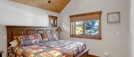 Un dormitorio con techo de madera, una cama grande con colcha colorida, una ventana con persianas y un cuadro enmarcado en la pared.