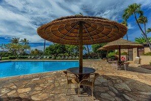 A pool area with a stone floor, a round table with chairs, and a large thatched umbrella.