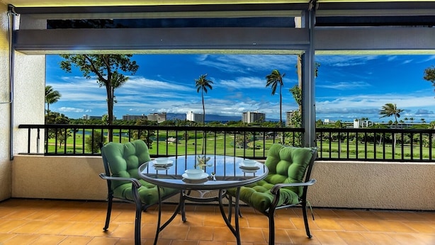 A balcony with a glass table and two green chairs, overlooking a golf course and city skyline.