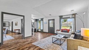 A spacious living area with a wooden floor, a sofa, a coffee table, and a lamp. There is a door leading to another room and a window with curtains. - 3 Bedroom Family Home in Lake Charles, La (Lake Charles)