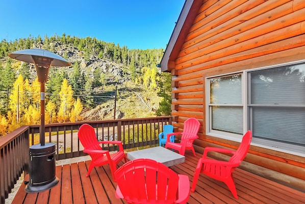 A wooden deck with a table and four chairs, a heater, and a view of a forested hillside.