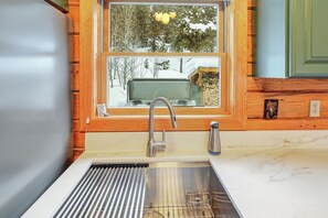 A kitchen with a stainless steel sink, wooden cabinetry, and a window with a view of trees and a shed.