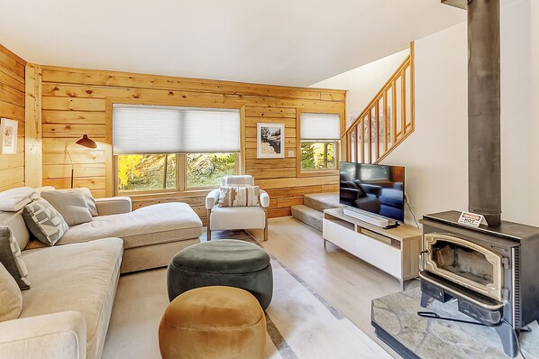 A cozy living room with a sofa, ottoman, TV, and a wood-burning stove.
