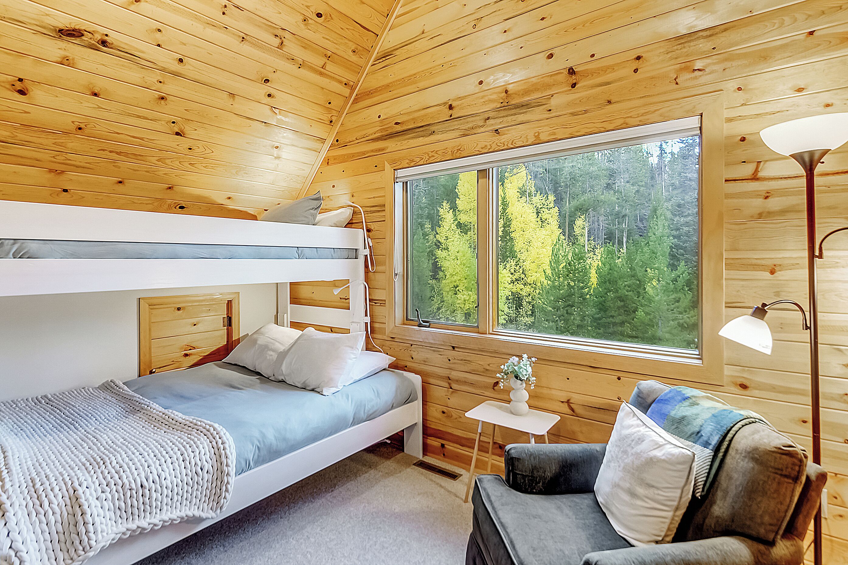 A wooden cabin bedroom with bunk beds, a small table, a chair, and a large window offering a forest view.