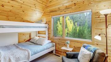 A wooden cabin bedroom with bunk beds, a small table, a chair, and a large window offering a forest view.