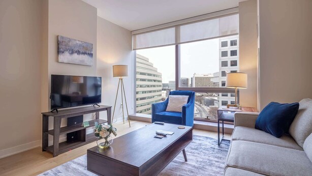 A modern living room with a flat-screen TV, a coffee table, a blue armchair, a beige sofa, and a large window offering a city view. - Modern Apt Downtown (Stamford, CT)