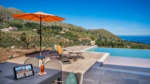 A poolside area with a view of the sea, a lounge chair, an umbrella, and a tablet displaying a travel theme. - Villa sulla Roccia, privacy e relax. (Sicilia)