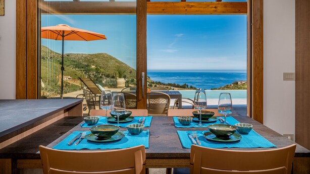 A dining table set with blue placemats, bowls, and wine glasses, overlooking a scenic coastal view. - Villa sulla Roccia, privacy e relax. (Sicilia)