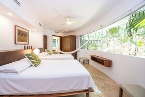 Habitación de hotel con dos camas, ventilador de techo, ventana grande con vista a la vegetación y un banco de madera.