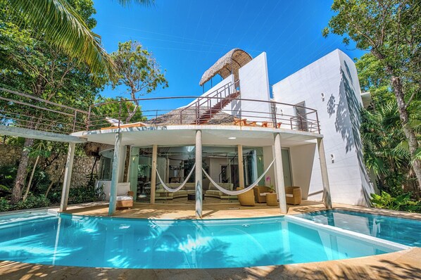 Una casa moderna con piscina, que cuenta con un porche con paredes de vidrio y una estructura de techo de paja.