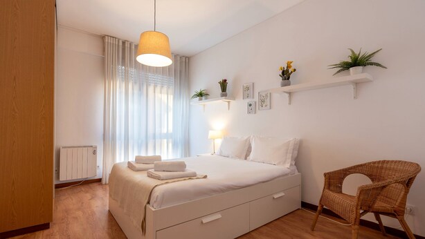 Ein Schlafzimmer mit einem großen Bett, einem Nachttisch mit Lampe, einem Sessel und einem Fenster mit Vorhängen.