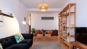 Ein modernes Wohnzimmer mit einer schwarzen Couch, zwei Korbstühlen, einem Holzregal, einem Flachbildfernseher und einer Pendelleuchte.