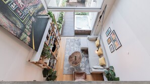 Un interior moderno con una escalera que lleva a un balcón, una zona de estar con cojines, y un cuadro abstracto grande en la pared.