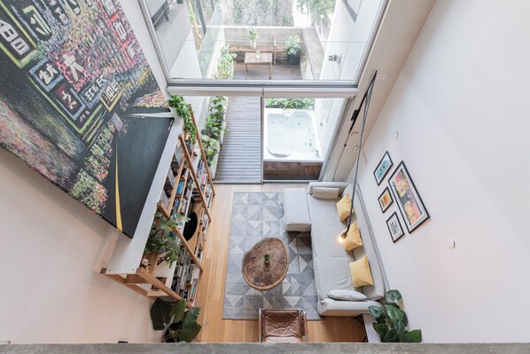 Et moderne interiør med en trappe op til en balkon, et siddeområde med puder, og et stort abstrakt maleri på væggen.