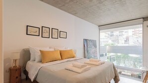 Un dormitorio con cama, mesita de noche, ventana que ofrece vistas a edificios y cuadros en la pared.