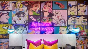 Un dormitorio con un colorido mural al estilo de cómic en la pared, un letrero de neón y una cama con almohadones amarillos y rosados.